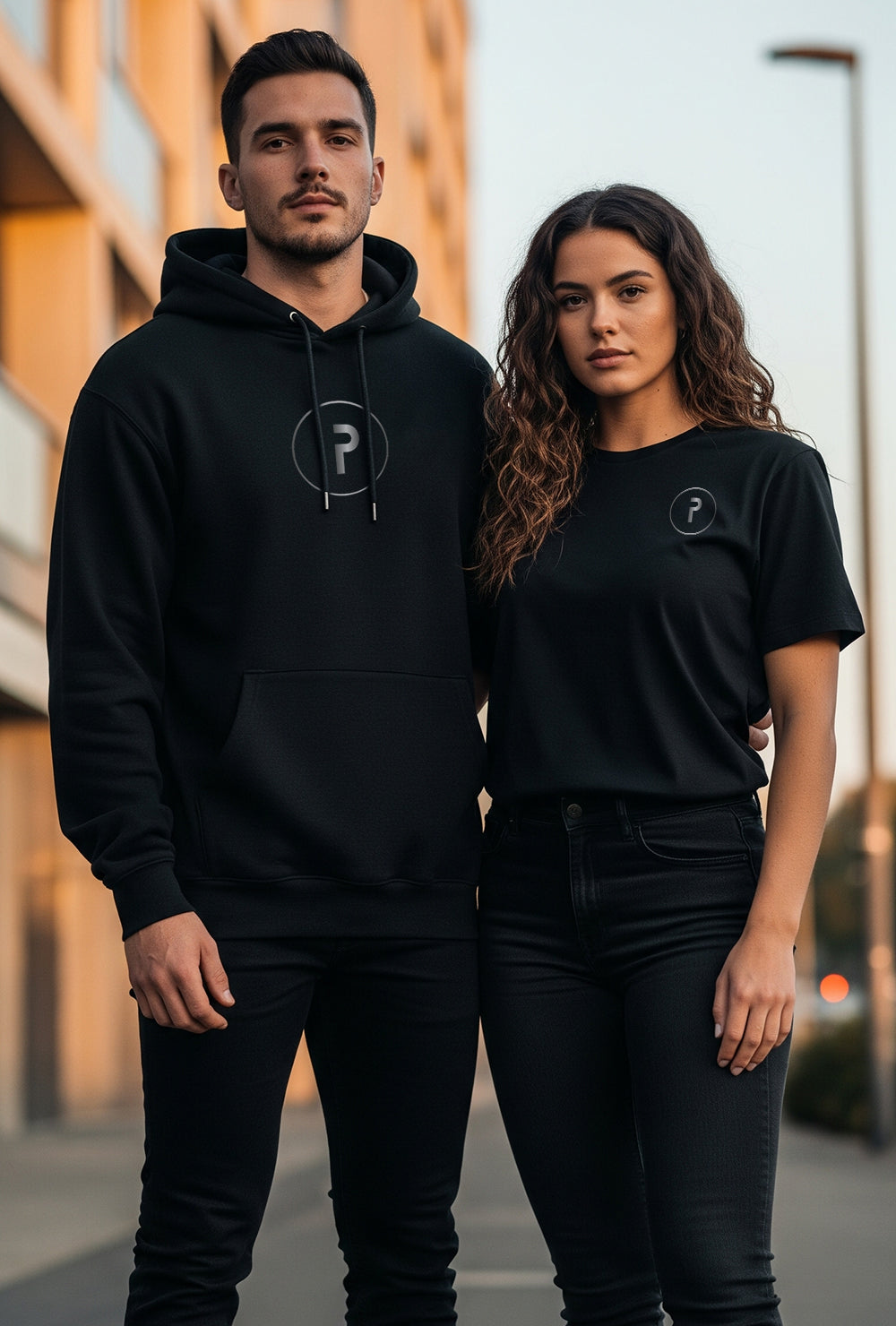 Two people wearing black hoodies and t-shirts with a logo, standing outdoors.